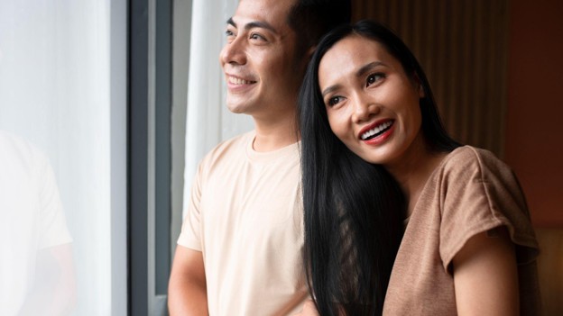 Pasangan tersenyum di dekat jendela dengan perempuan menyender di bahu laki-laki menggambarkan kebutuhan rasa aman dalam hubungan dan reassurance seeking