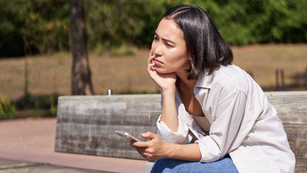 Ilustrasi seorang perempuan yang duduk sendirian di bangku taman sambil memegang ponsel dengan ekspresi murung. Gambar ini menggambarkan dampak toxic positivity, ketika seseorang merasa harus selalu terlihat kuat dan positif meskipun sedang menghadapi tekanan emosional, stres, atau kekecewaan. Mengakui dan menerima emosi yang tidak nyaman merupakan langkah penting dalam menjaga kesehatan mental dan mengelola emosi secara sehat.